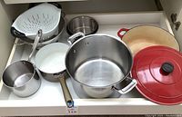 Multiple cookware items displayed in cabinet drawer including red Dutch oven, stainless steel pots, frying pan, and colanders