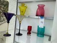 Full view of the colorful glass items on glass shelves, showing purple and frosted martini glasses, yellow vase, and several votive holders in red and turquoise.