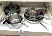 drawer view showing a stack of various stainless steel skillets with grey silicone protectors and a saucepan at front left