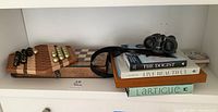 Shelf view showing exterior of fold-out board game with Chinese checkers marbles, stack of books, binoculars and stone coasters