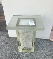 Front view of square glass side table showing textured clear glass top, blocky base with central textured insert