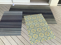 Three outdoor rugs displayed on deck: large striped rug at back, smaller striped rug at left, patterned rug at right.