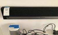Front view of black Sonos soundbar mounted on wall with seller tag, and white Sonos Play:1 speaker below, cables visible