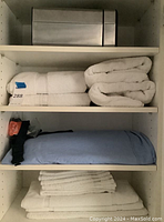 Shelving unit view: top shelf holds a stainless steel waste bin; second shelf shows four folded white towels; third shelf contains a light blue pillow or pillowcase bundle with black strap; bottom shelf features a stack of white hand towels atop a white bath mat.