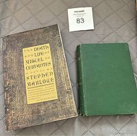 Photo shows two books placed side by side on a quilted surface. The left book is The Death and Life of Miguel de Cervantes with a decorated cover. The right book is older, with a plain green cloth cover.
