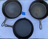 Top view of three cast iron frying pans with handles visible, arranged side by side on a white cloth. One has hammered edge and another is marked Wagner Ware.