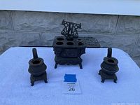 Set of three cast iron toy stoves displayed on a white cloth against a textured stone background. Central large stove has six burner holes, ornate sides, and a side shelf. Two smaller stoves flank it, each with a single burner opening and a handle.