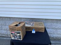 Two cardboard boxes containing Coleman camping equipment placed on the ground outside: one with Coleman lantern, one with 2 burner stove.