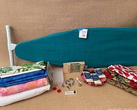 Wide view of sewing supplies lot including fabric, sewing machine parts, hooks, bias tape, and teal ironing board