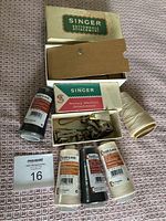 Vintage Singer buttonhole attachment in original box with additional Singer sewing machine attachments and various Threadi-Lock spools of thread in white, beige, and black.