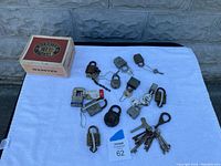 Mixed lot of vintage keys and various padlocks spread on white cloth background, including some attached keys and keychains.