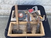 Photo showing two C-clamps, two woodworking planes, hatchet, measuring tapes inside a wooden box.
