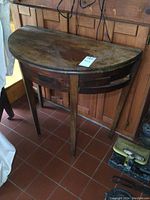 Photo of antique games table in half-round folded position against wood paneled wall, showing aged wooden surface and four tapered legs.
