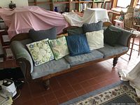 Full view of antique Victorian-style upholstered settee with six throw cushions on seat, showing overall condition and setting in room with tile flooring and covered furniture in background.