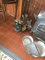 Full view of fireplace tools including poker sets on stands, bellows, and dust pans with shovels arranged on tiled floor near wood stove.