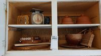 Two shelves of assorted kitchen items in a wooden cabinet, showing covered pottery butter dishes, wooden bowls, cheese boards, and a kitchen timer and scale.