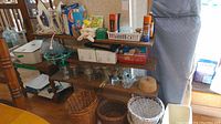 Three shelves with assorted laundry and cleaning supplies, two irons, baskets, and jars; an ironing board leaning against the wall