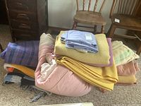 View of stacked wool blankets, pillowcases, and a pink comforter on carpeted floor with wooden chairs in background.