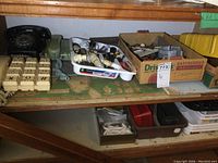 Shelf with black rotary dial phone, green corded phone, beige cutlery trays, plastic hardware tray, and cardboard box with miscellaneous small metal parts and handles.