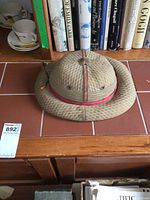 Original fabric covered pith helmet from India with quilted appearance and red band, placed on tiled surface.