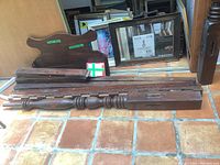 Disassembled wood bed frame components including turned posts and side rails stacked on tiled floor.