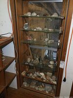 Wide view of wooden cabinet with five glass shelves displaying a variety of rock specimens and some empty bottles, with a wooden shelving unit next to it.