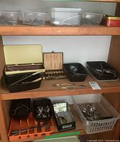 Shelf with multiple containers holding various silverware and utensils including a yellow-lined case with Apostle spoons.