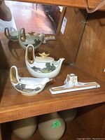 Photo showing the teapot, creamer, and pen holder on a wooden shelf with their painted landscape and gold details.