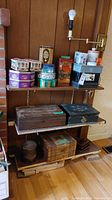 Front view of a shelf with multiple tin containers on the top shelf and wooden boxes on two lower shelves against a wooden wall.