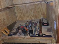 Multiple vintage hand tools including saws, hand drills, hammers, wrenches, and a carpenter's square laid out on a wooden shelf inside a plywood enclosure.