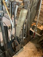 Photo showing a range of vintage yard tools including shovels, pickaxe, post hole digger, and axe wedge propped against wall in garage.