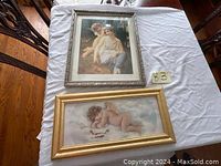 Two framed angel pictures displayed on a white cloth with a tag number 83. One silver ornate frame with a seated angel picture, one gold frame with a sleeping angel picture.