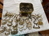 Photo of multiple antique brass drawer handles of different styles and sizes arranged on a white cloth with some handles grouped and others loose, including a brass container holding additional handles.