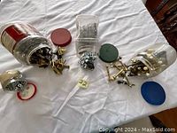 Four plastic jars with lids in different colors, containing brass window locks, door stoppers, screws, and washers spread on a white surface.