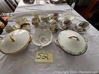 Photo showing a porcelain set including two lidded soup containers, several creamers, cups, saucers, and a white glass basket on a table.