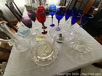 Photo showing various colored wine glasses including red, blue, purple, and teal along with clear glass and crystal items like a decanter and bowls on a white tablecloth.