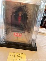 Full view of boxed collector doll in protective display case on black base with ornate window and collector series labeling.