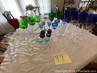 Wide view of varied crystal and glass cups in multiple colors placed on a white tablecloth showing size and quantity.
