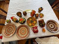 Top-view photo showing two large ceramic plates, a medium tray, and numerous small ceramic decorative jars, pots, pitchers with floral folk art patterns in earthy colors.