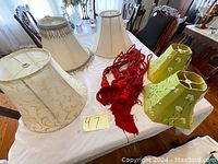 Lot includes six lamp shades of various styles in white, off-white, and green fabric, and two red curtain tiebacks with tassels, displayed on a white tablecloth indoors.