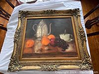 Large still life painting featuring a silver coffee pot, fruits, and a cup, displayed on a table with an ornate gold frame.