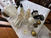 Lot of one large clear glass hanging lamp, ten diverse glass lamp shades, and one small black and brass wall lamp base arranged on a white cloth surface.