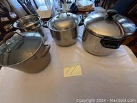 Four large pots with lids displayed on a table. Pots are of different styles and finishes, including copper-colored and stainless steel.