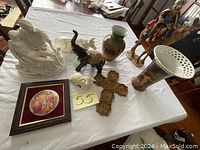 Photo shows a table with a religious figurine statue, a carved cross, multiple porcelain vases including one with an open lattice top, animal figurines (elephant, rabbit, horse), framed religious artwork, and a multi-figure religious statue.