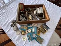 Box containing various metal hinges, some still in packaging, and other hardware items on a white surface.