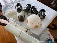 Table top view of five black metal show lamps, two glass shades, and one large rectangular light fixture arranged on a white tablecloth.
