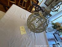 View of the entire vintage metal outdoor hanging lamp fixture placed on a table with number 87 tag nearby. Shows scrollwork pattern and hanging loop on top.