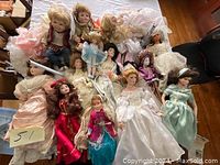 Photo showing 14 vintage porcelain dolls in assorted elaborate dresses and various sizes, arranged on a bedspread.