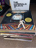 Stack of vinyl LPs including visible albums: The Imperials 'Believe It' and Saturday Night Fever soundtrack.