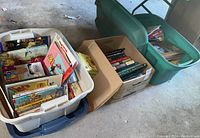 Three bins filled with assorted books including children's books, nutrition, religion, and adoption themes. Bins are one green plastic tub, two white plastic tubs with lids, and one cardboard box.